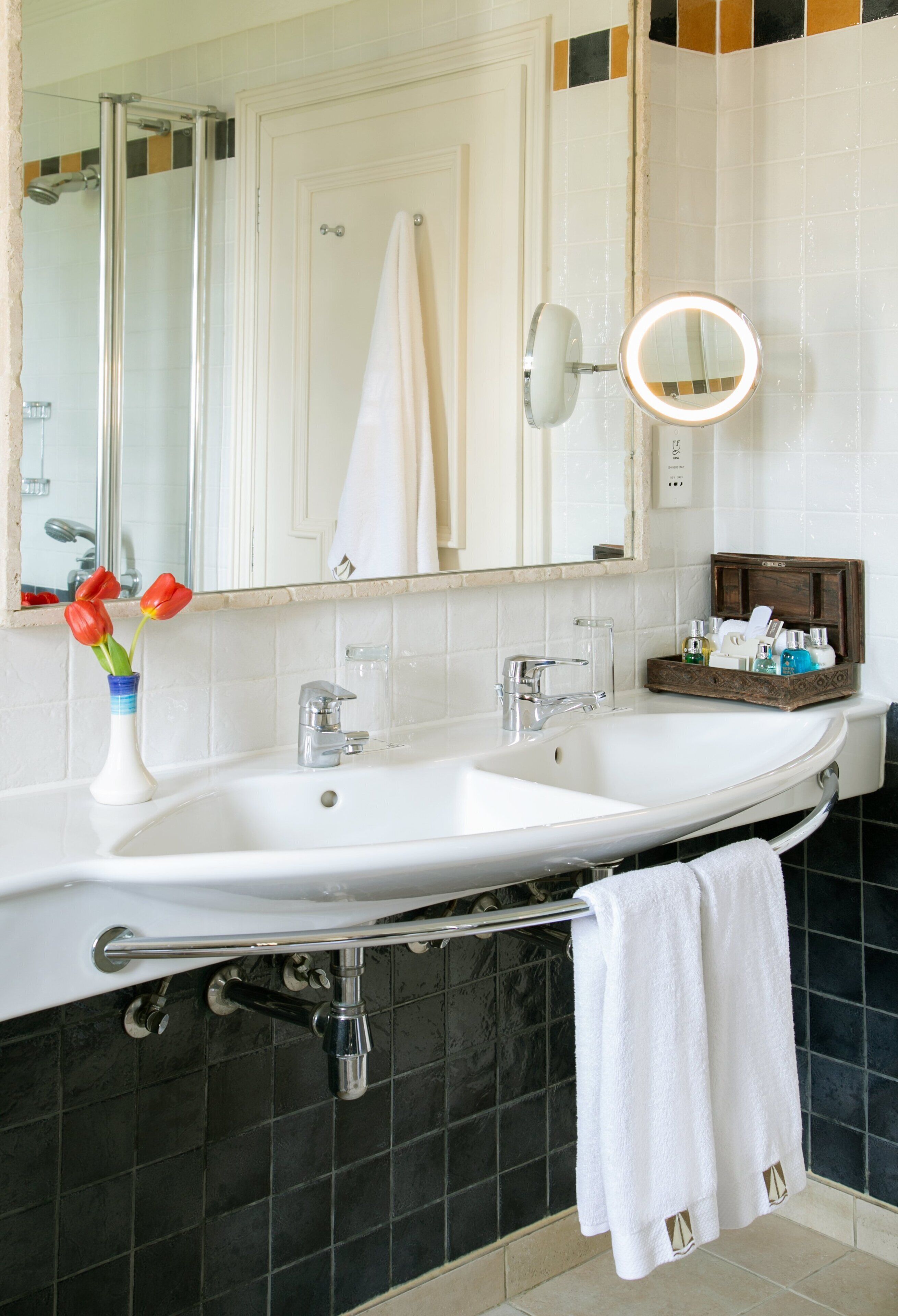 A bathroom with a large white sink, a mirror with a light, a towel rack, and a vase with flowers.