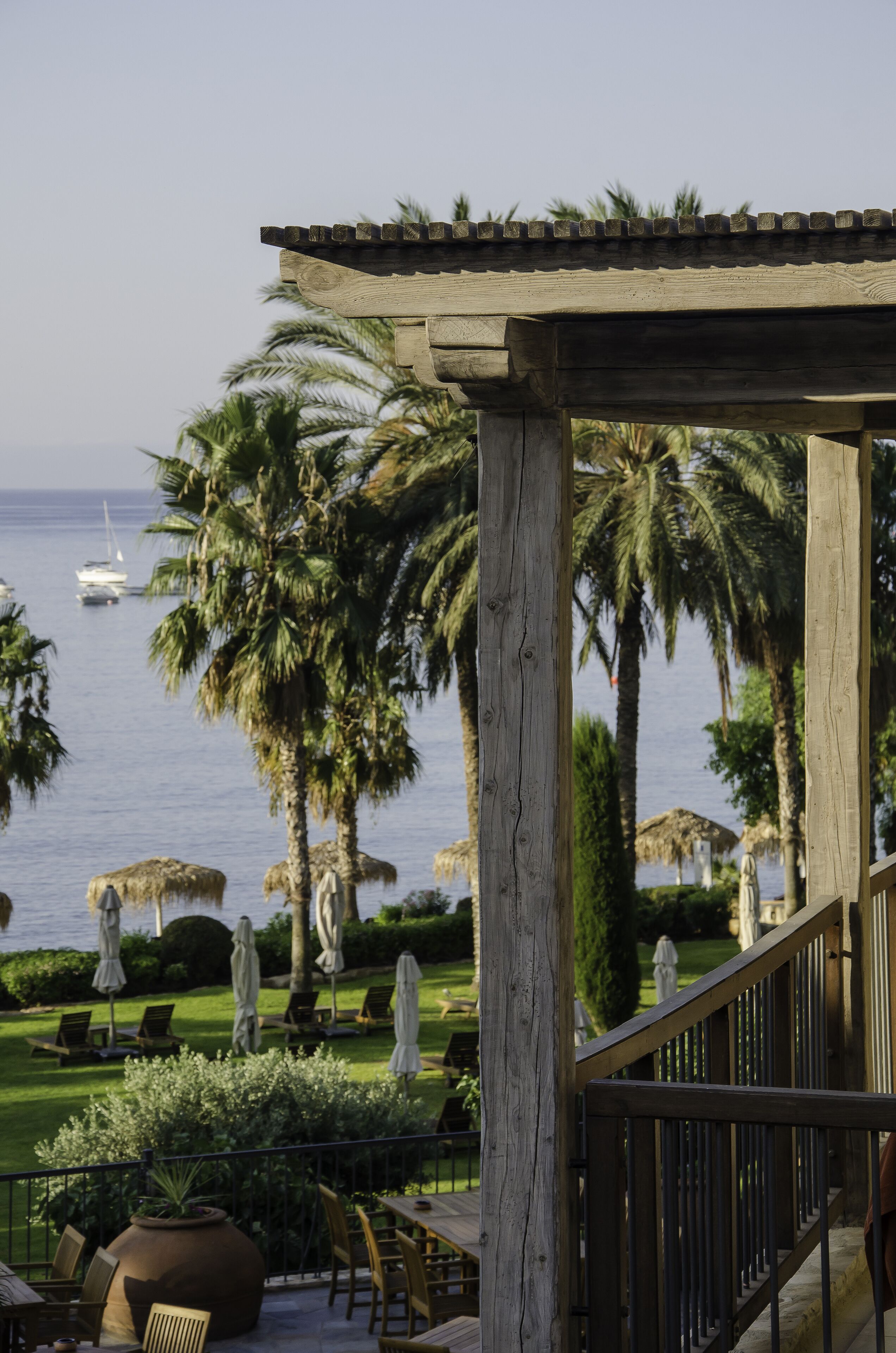 Una pérgola de madera con vistas a una playa con palmeras, sillas de descanso y vista al mar.