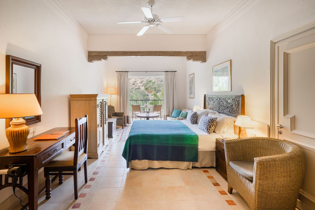 A hotel room with a bed, a desk with a lamp, a chair, a balcony with a table and chairs, and a ceiling fan.