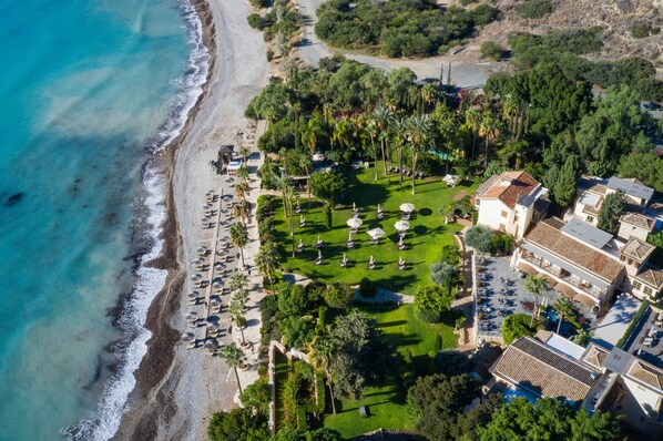 An aerial view of a coastal resort with a beach, palm trees, and a parking area. - Columbia Beach Resort (Pissouri)