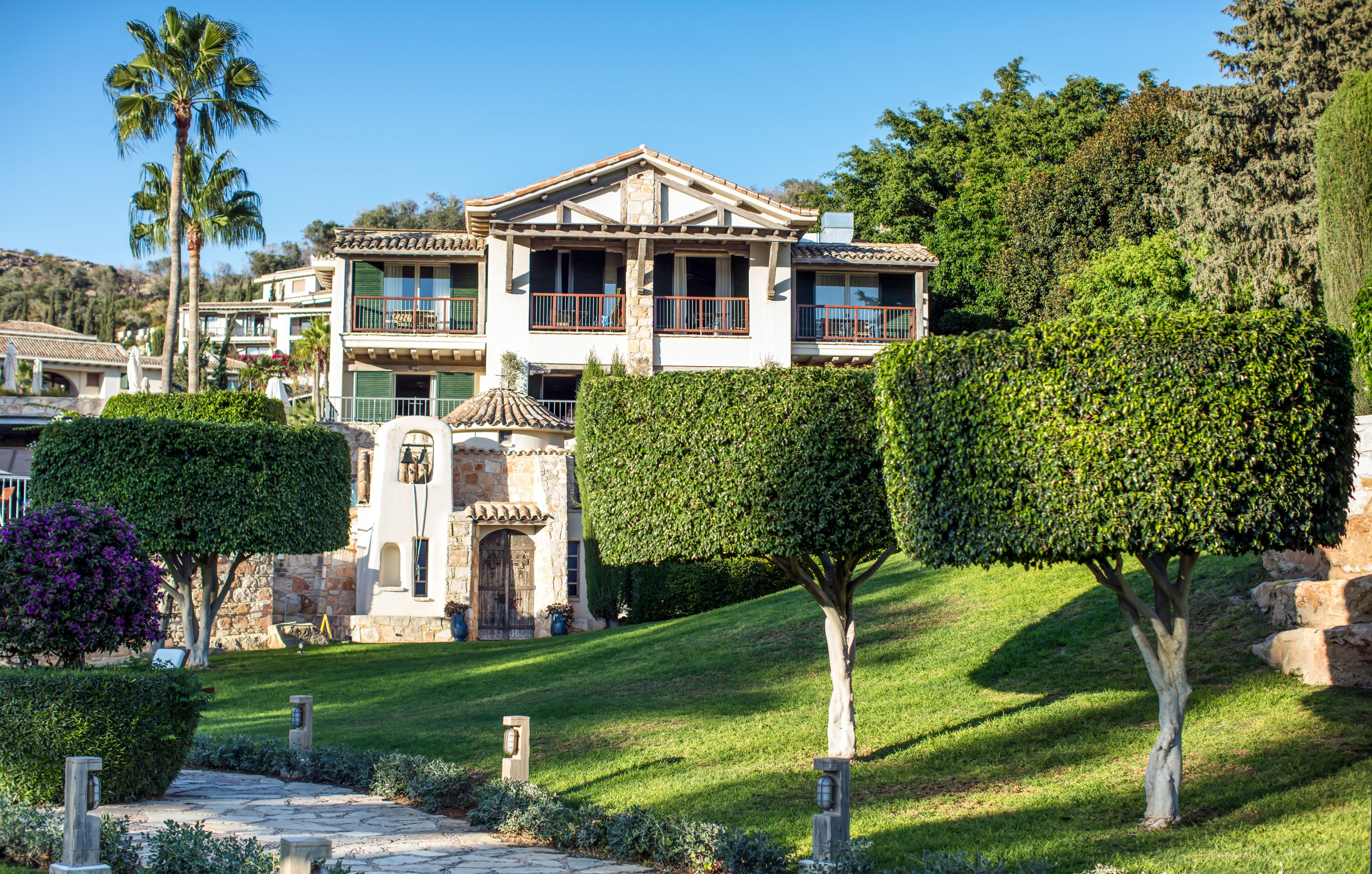 Una casa grande de dos pisos con fachada de piedra, varios balcones y un jardín cuidado con setos podados y una palmera.