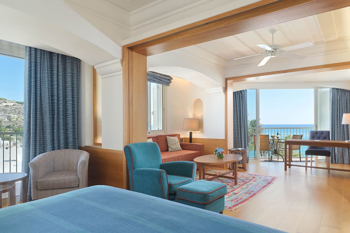 A hotel room with a bed, two armchairs, a sofa, a dining table, and a balcony with a view of the sea.