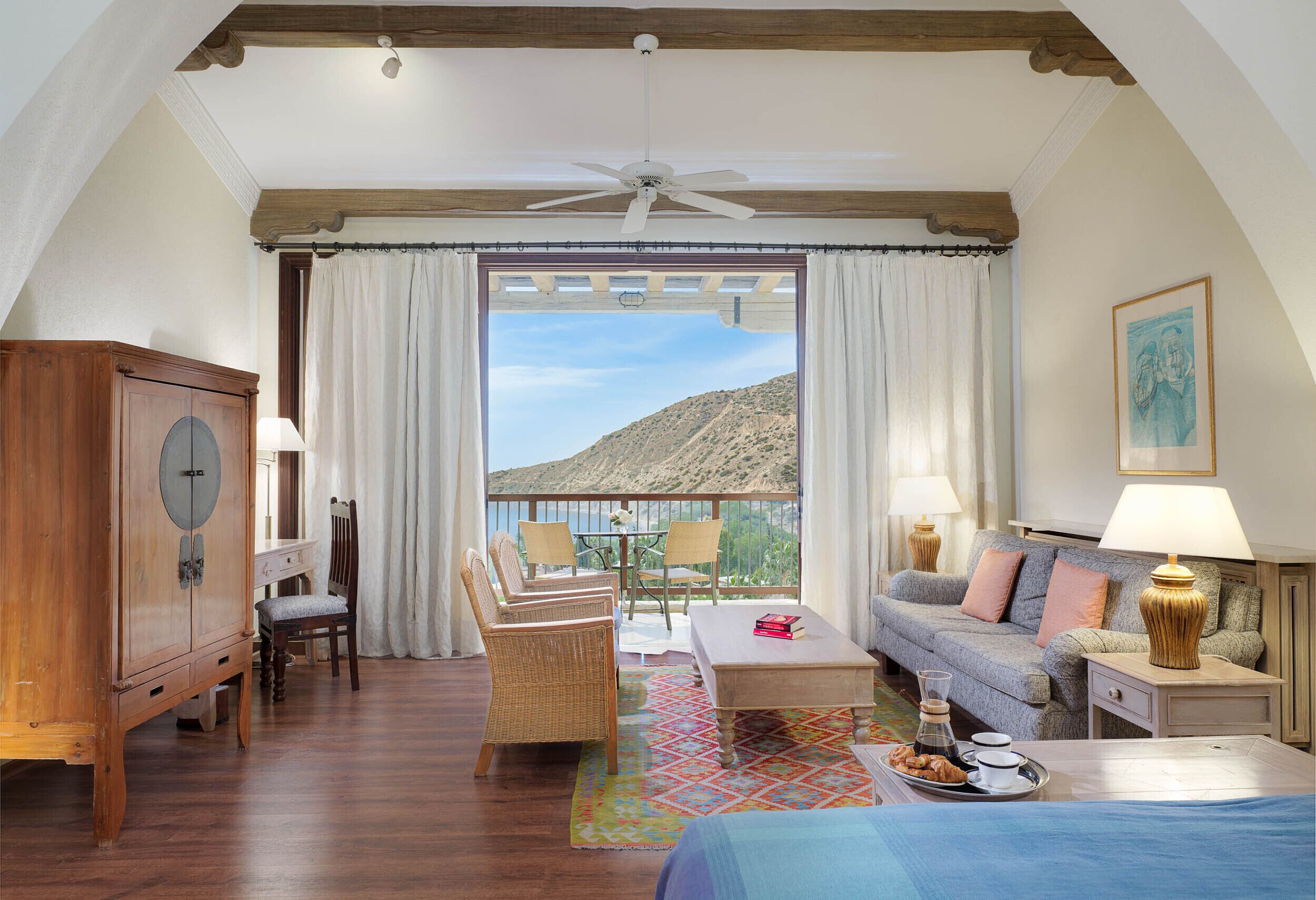 A spacious room with a large window, a ceiling fan, a wooden cabinet, a sofa, a dining table with chairs, and a bed.