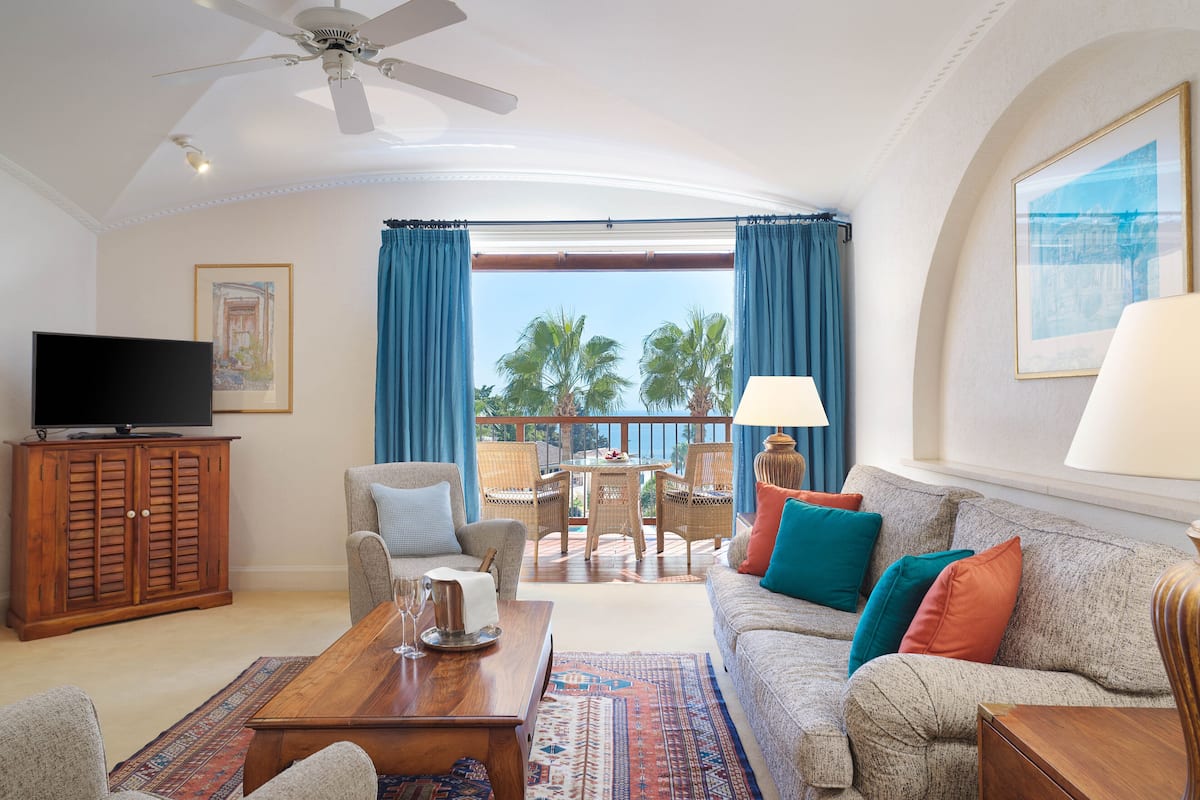 A living room with a sofa, chairs, a coffee table, a TV, and a balcony with a view of palm trees.