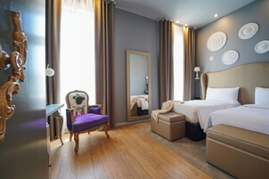 A hotel room with a wooden floor, a bed with white linens, a chair with a purple cushion, a small table, and a mirror on the wall.