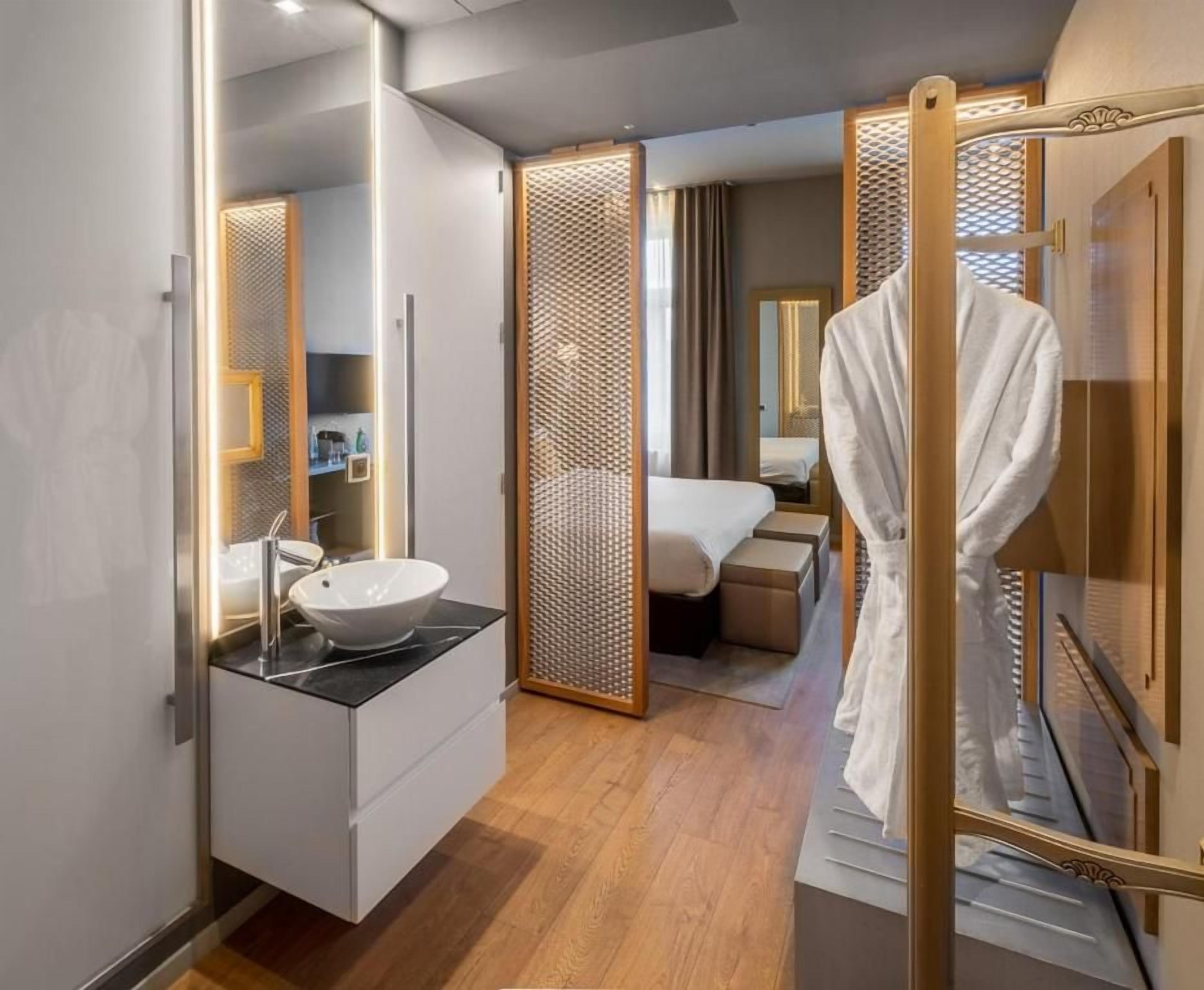 A modern hotel room with a wooden floor, a white bathroom with a round sink, a bed with a white duvet, and a robe hanging on a door.