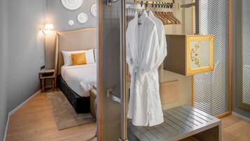 A modern hotel room with a glass partition separating the bedroom and bathroom. The room features a bed with white bedding, a bedside table, and a wall-mounted lamp.