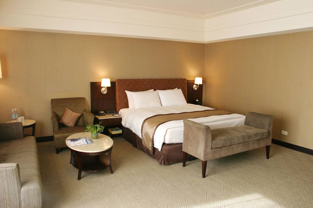 Une chambre d’hôtel avec un grand lit, deux fauteuils, une petite table avec une plante et une table de chevet avec une lampe.