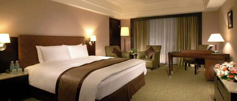 Habitación de hotel con una cama grande, dos sillones, un escritorio y una mesa de comedor. Hay lámparas a ambos lados de la cama y una ventana con cortinas.