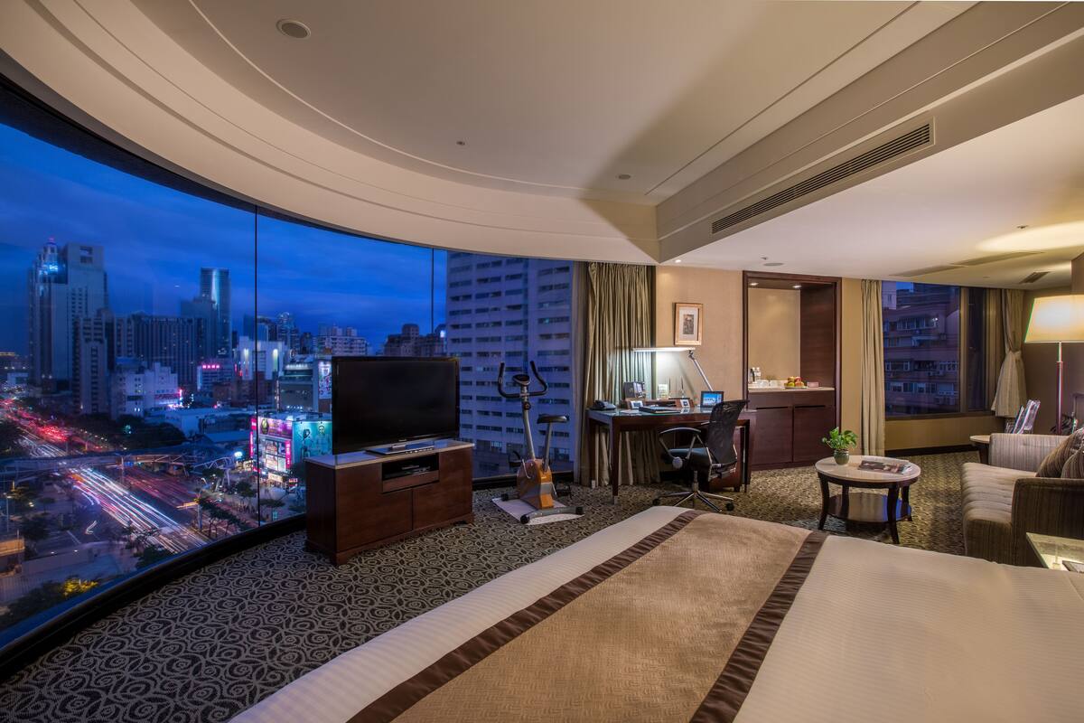 Une chambre d’hôtel avec un grand lit, un bureau, une télévision et une vue sur le paysage urbain illuminé la nuit.