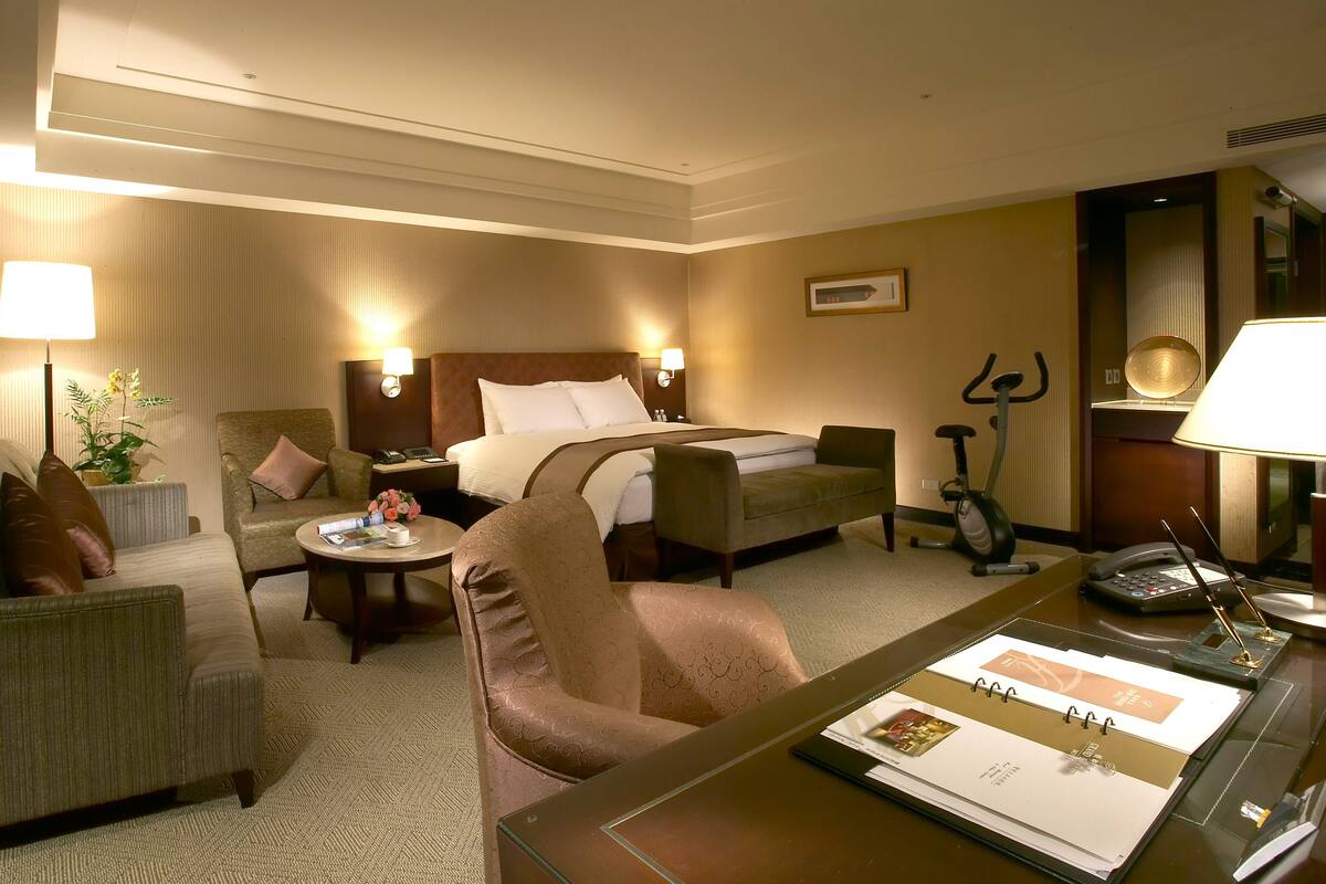 Une chambre d’hôtel comprenant un lit, deux fauteuils, un bureau avec une lampe, un vélo d’appartement et une petite table avec une plante et un magazine.