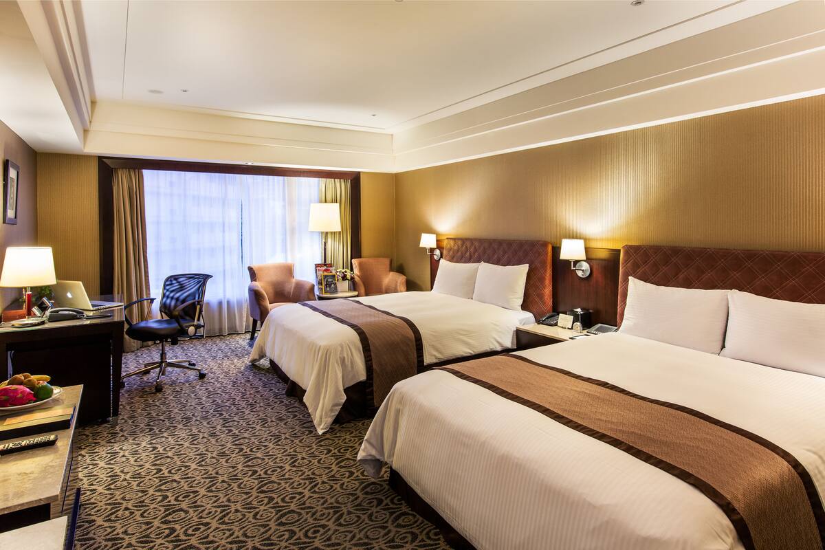 Une chambre d’hôtel avec deux lits, un bureau avec un ordinateur portable, une chaise et une grande fenêtre avec des rideaux.