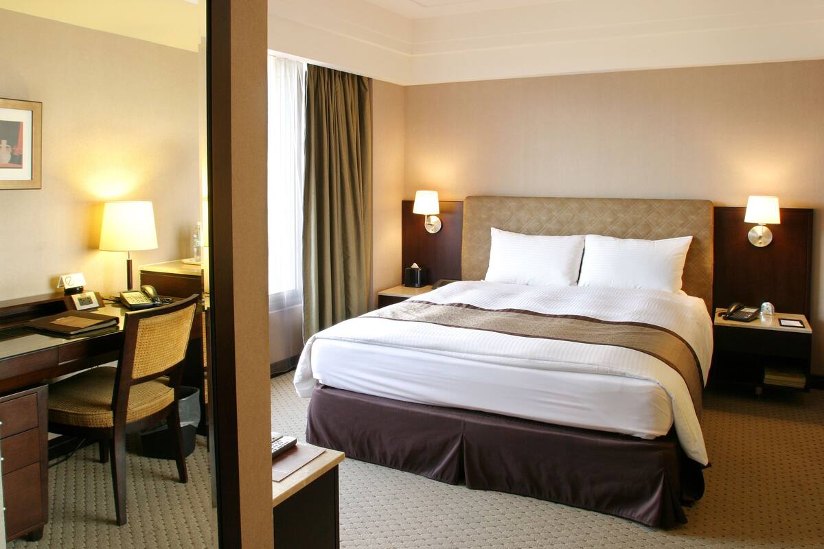 Une chambre d’hôtel avec un grand lit, deux tables de chevet avec des lampes, un bureau avec une chaise et une fenêtre avec des rideaux.