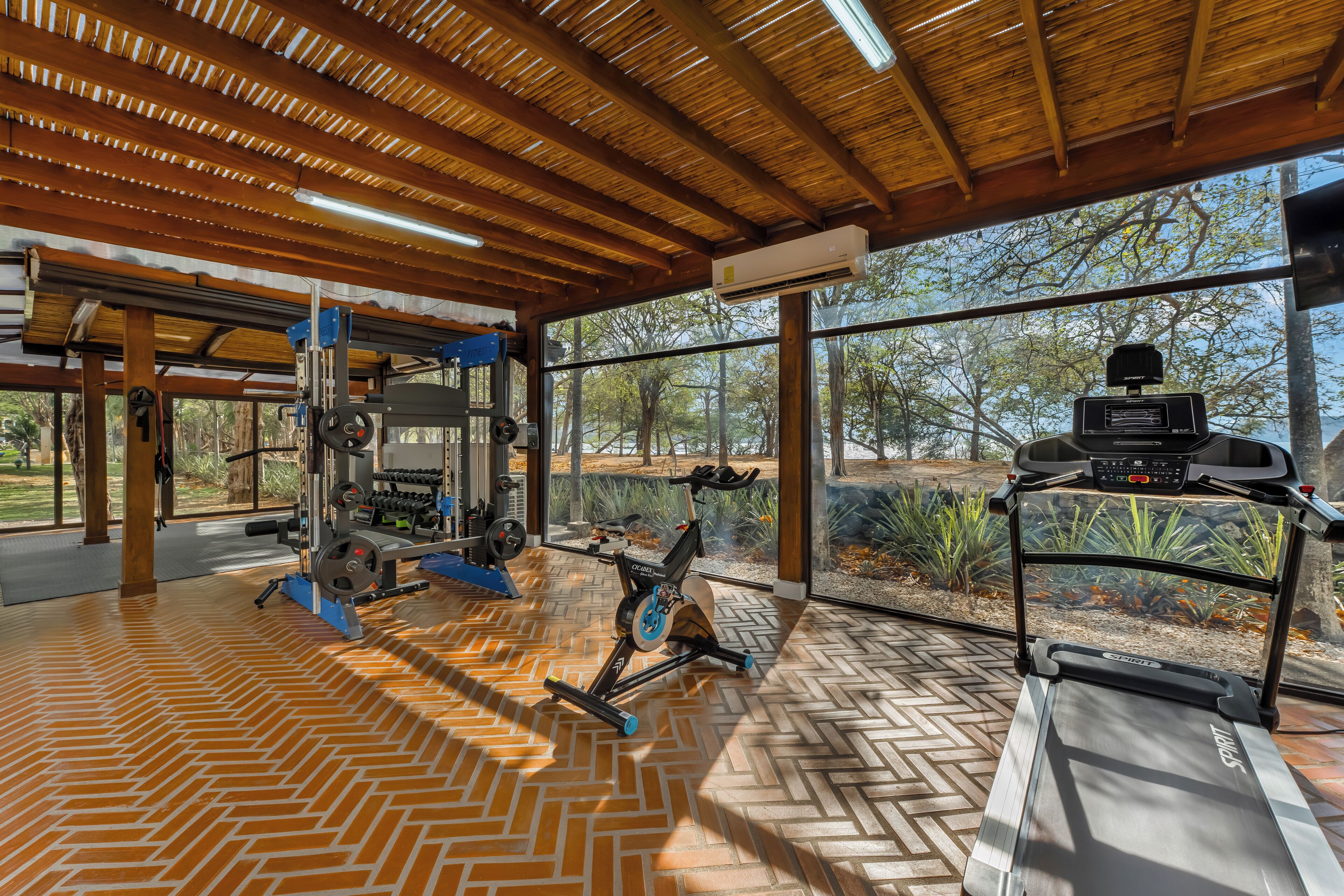 Une salle de sport équipée de matériel d’exercice, dotée d’un sol à motifs en chevrons et de grandes fenêtres offrant une vue sur les arbres.