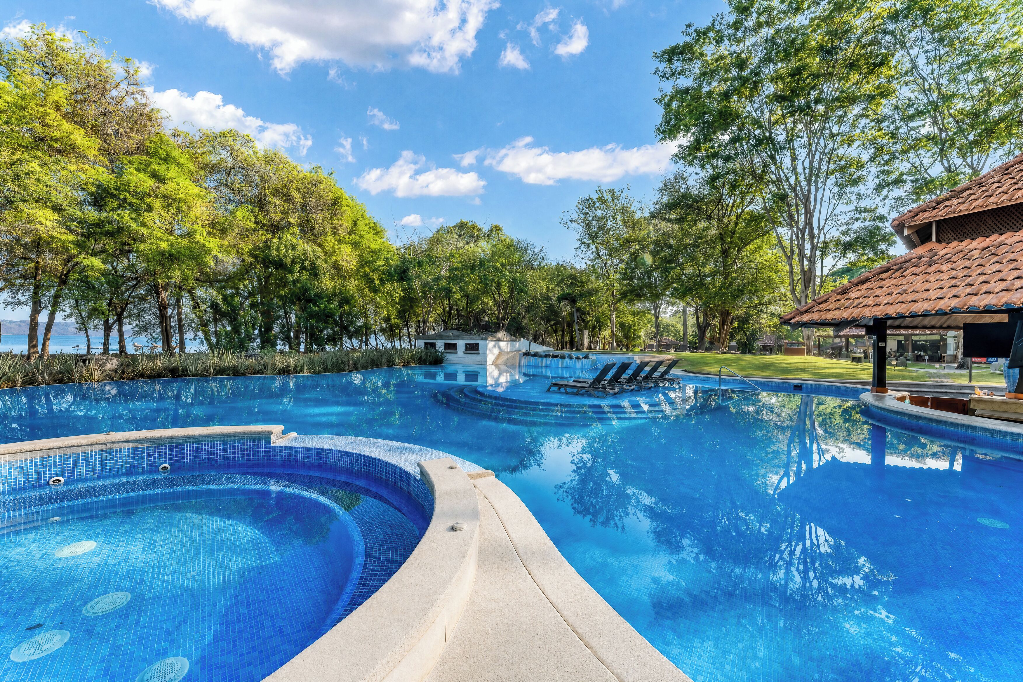 A swimming pool with clear blue water, surrounded by trees and a building with a tiled roof.