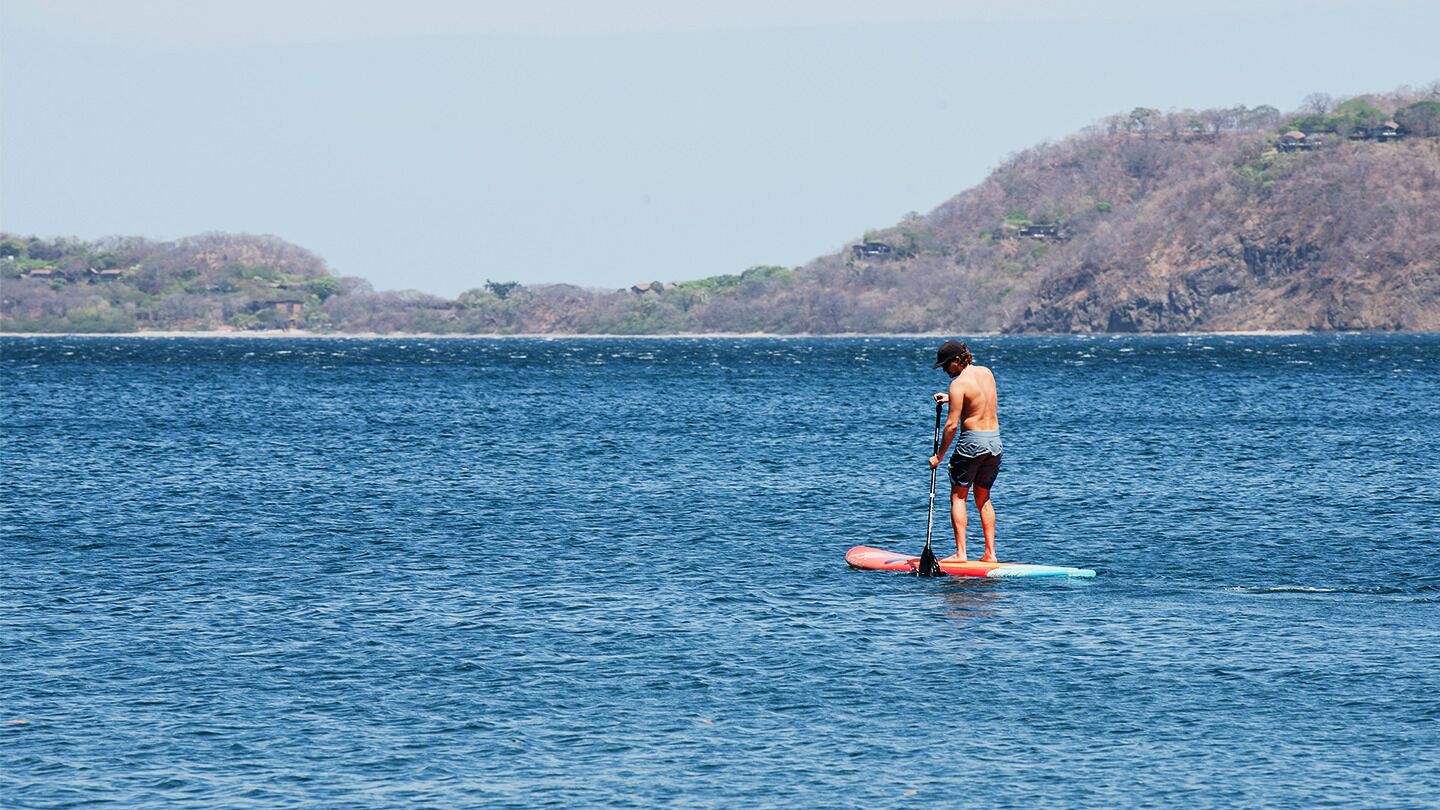 A person paddleboarding on a calm sea with a hilly landscape in the background.