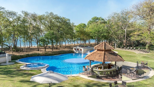 A swimming pool with a tropical gazebo, surrounded by trees and a view of the sea.