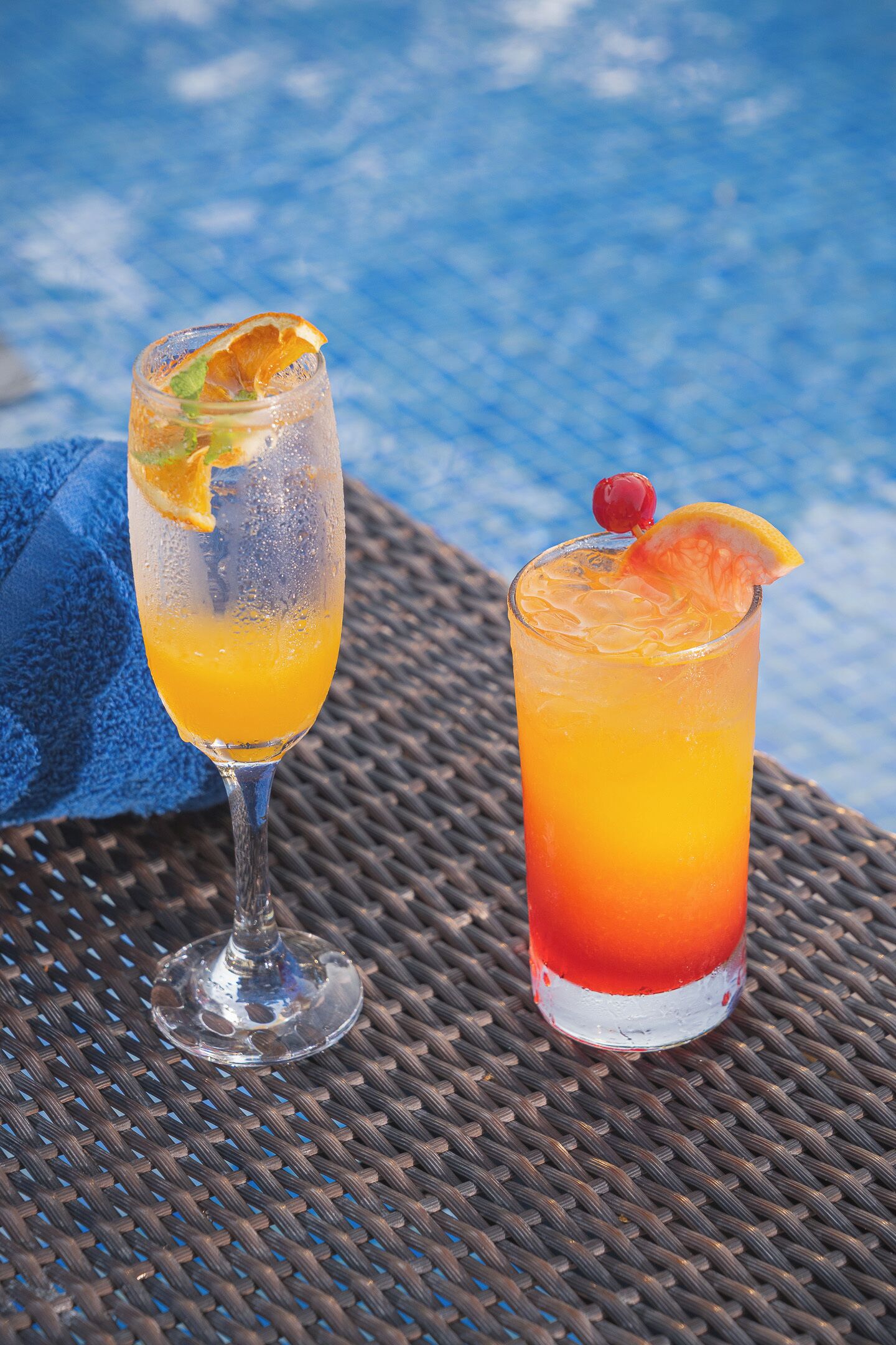 Two glasses of orange beverage on a woven table with a blue towel and a pool in the background.
