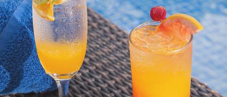 Two glasses of orange beverage on a woven table with a blue towel and a pool in the background.