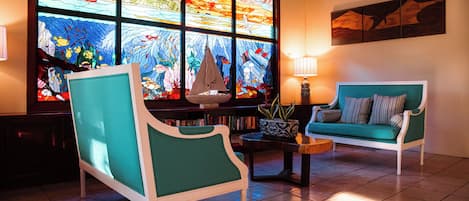 Una habitación con un sofá verde, un sillón blanco, una mesita y un amplio ventanal con vitrales.