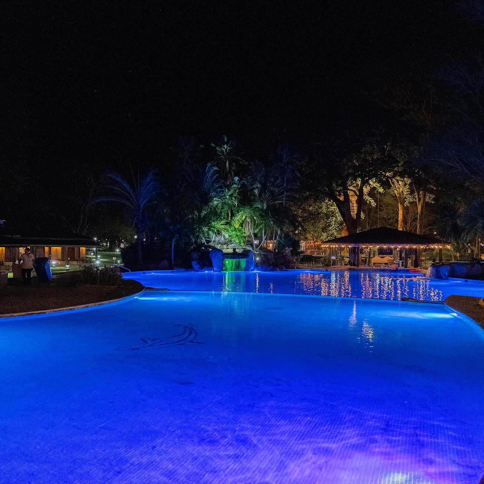 A nighttime swimming pool with illuminated edges and a waterfall feature.
