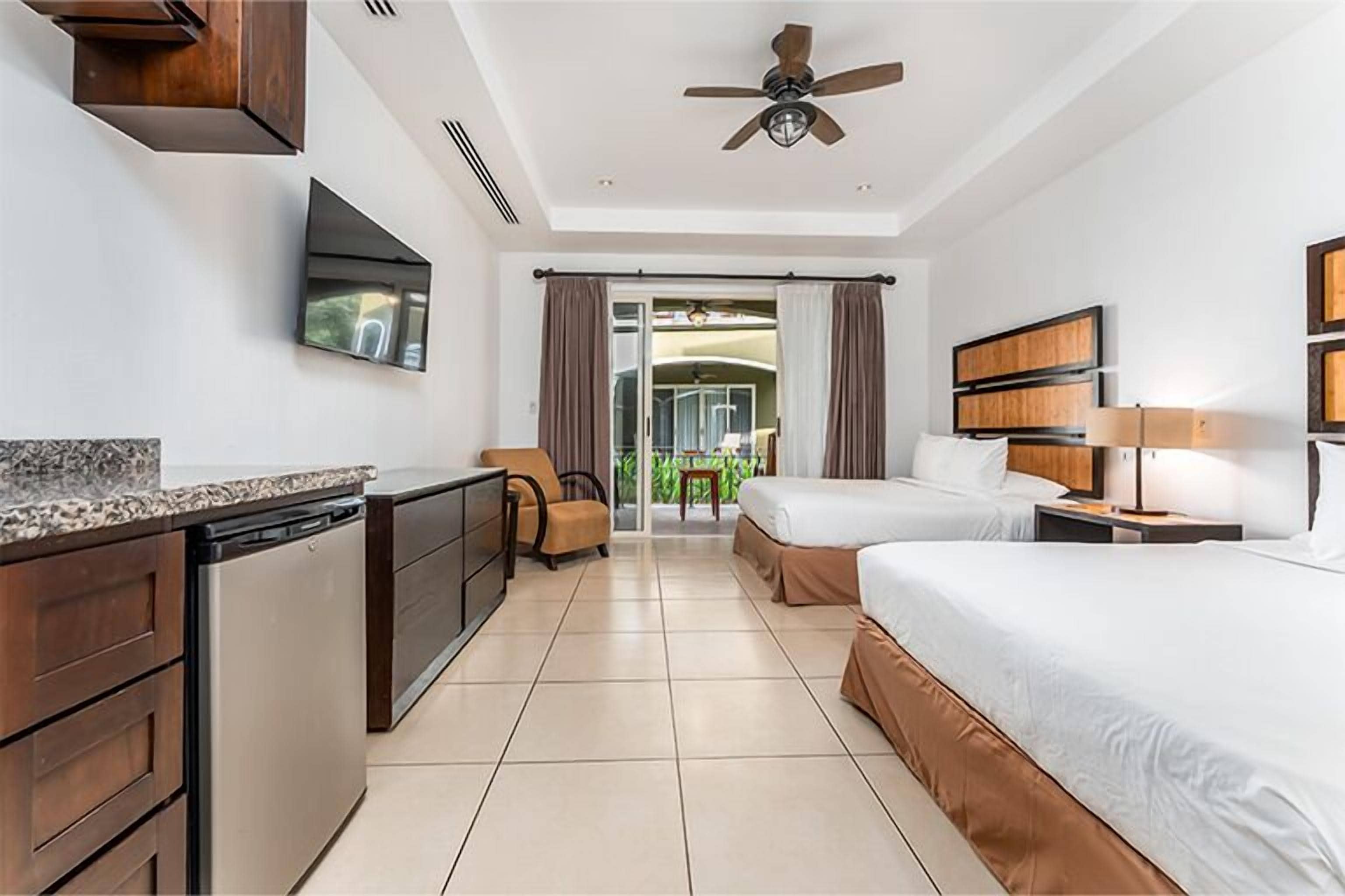 A hotel room with a kitchenette, two beds, a TV, and a ceiling fan.