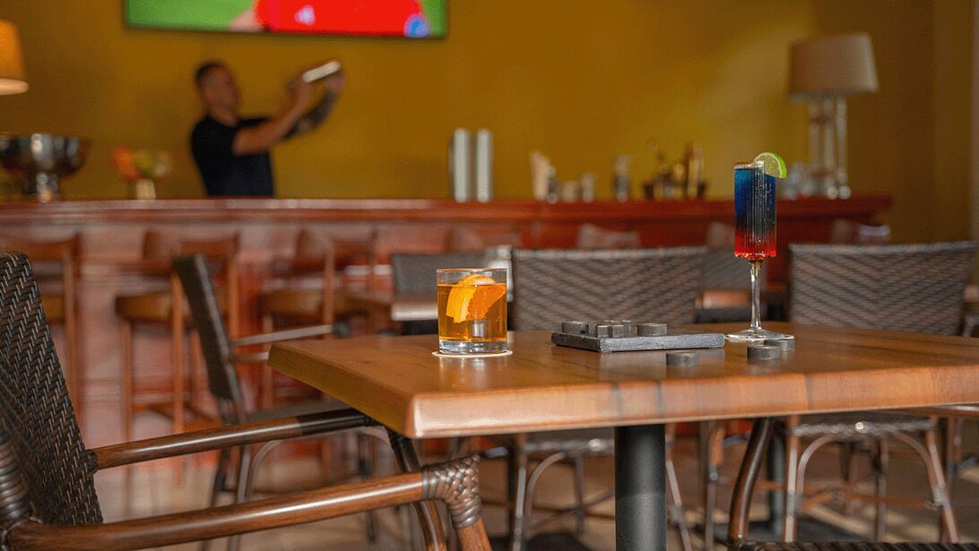 Un coin bar avec deux boissons sur une table, un téléviseur en arrière-plan et une personne derrière le comptoir.
