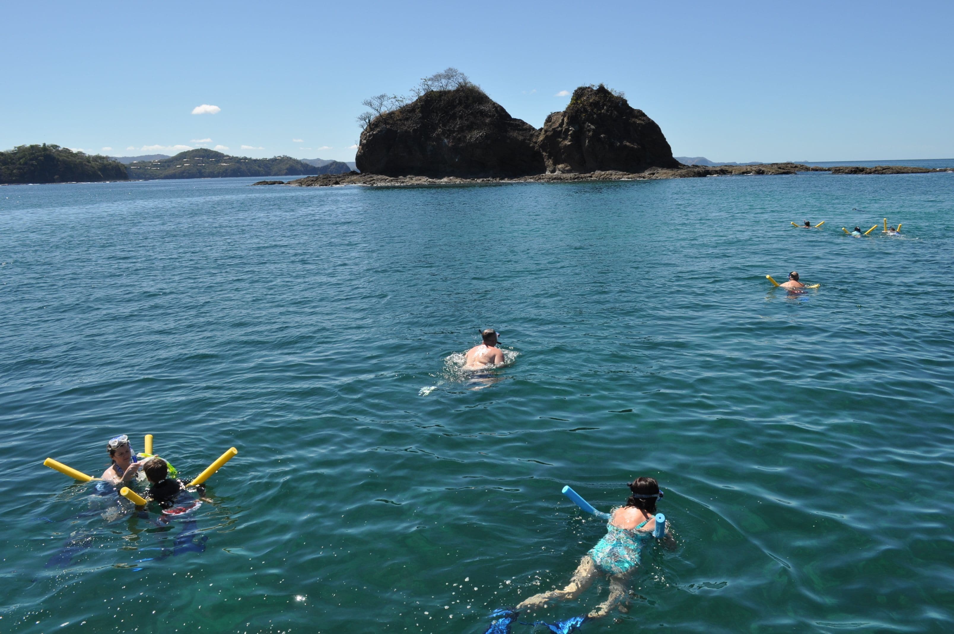 Un grupo de personas nadando y flotando en un mar tranquilo con afloramientos rocosos al fondo.