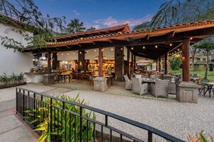 Un espacio exterior para comer con un porche cubierto, disposición de mesas y sillas, y vista del edificio contiguo.