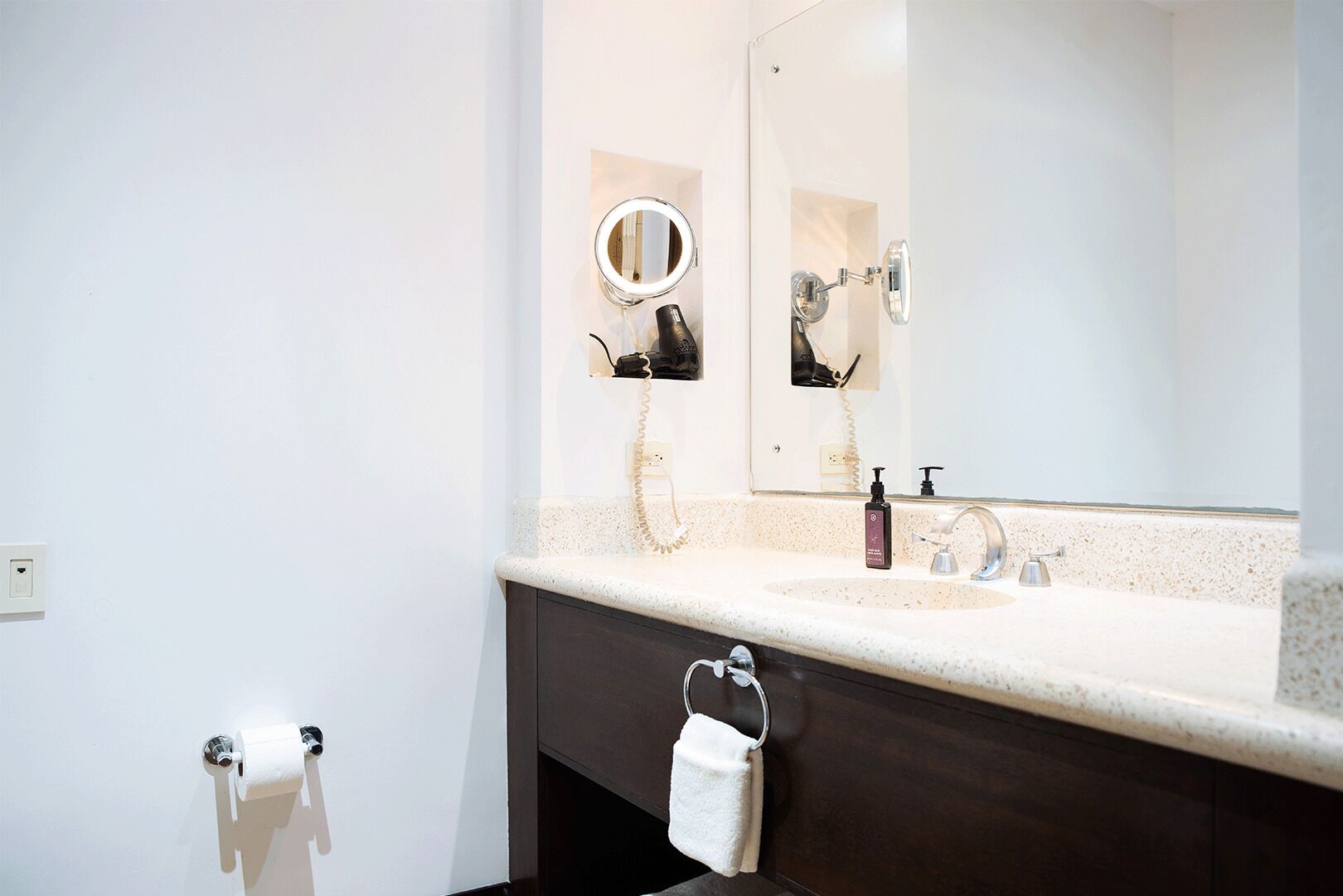 A bathroom with a white countertop, a sink, a mirror with a round mirror, a towel rack, and a toilet paper holder.