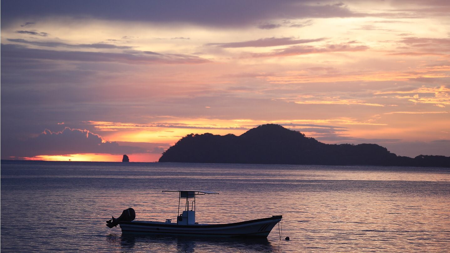 Un bateau sur une eau calme, avec un coucher de soleil et une île lointaine en arrière-plan.