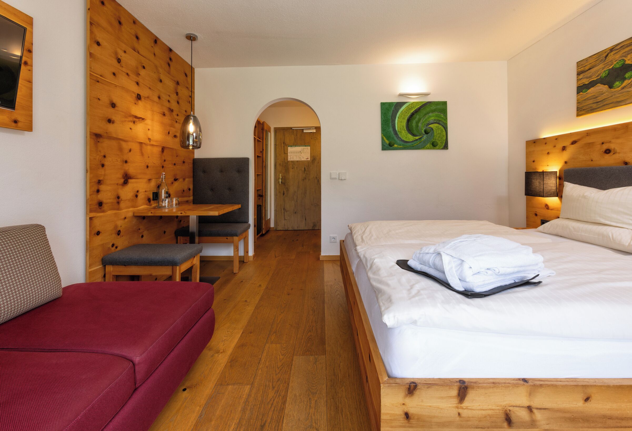Une chambre d’hôtel avec un mur en bois, un lit avec du linge de maison blanc, un canapé rouge, un bureau avec une chaise et un tableau accroché au mur.