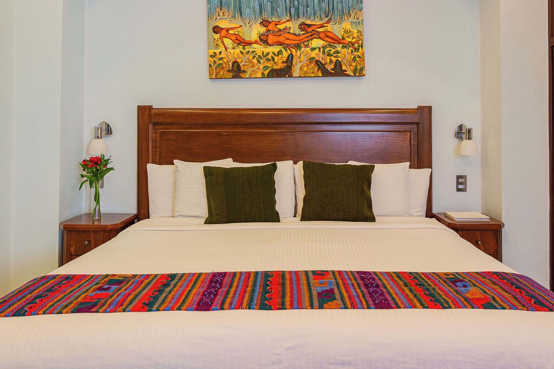 Ein Hotelzimmer mit einem hölzernen Kopfteil, einem großen Bett mit weißer Bettwäsche und grünen Kissen, einer farbenfrohen Decke, einem Gemälde über dem Bett und Nachttischlampen.