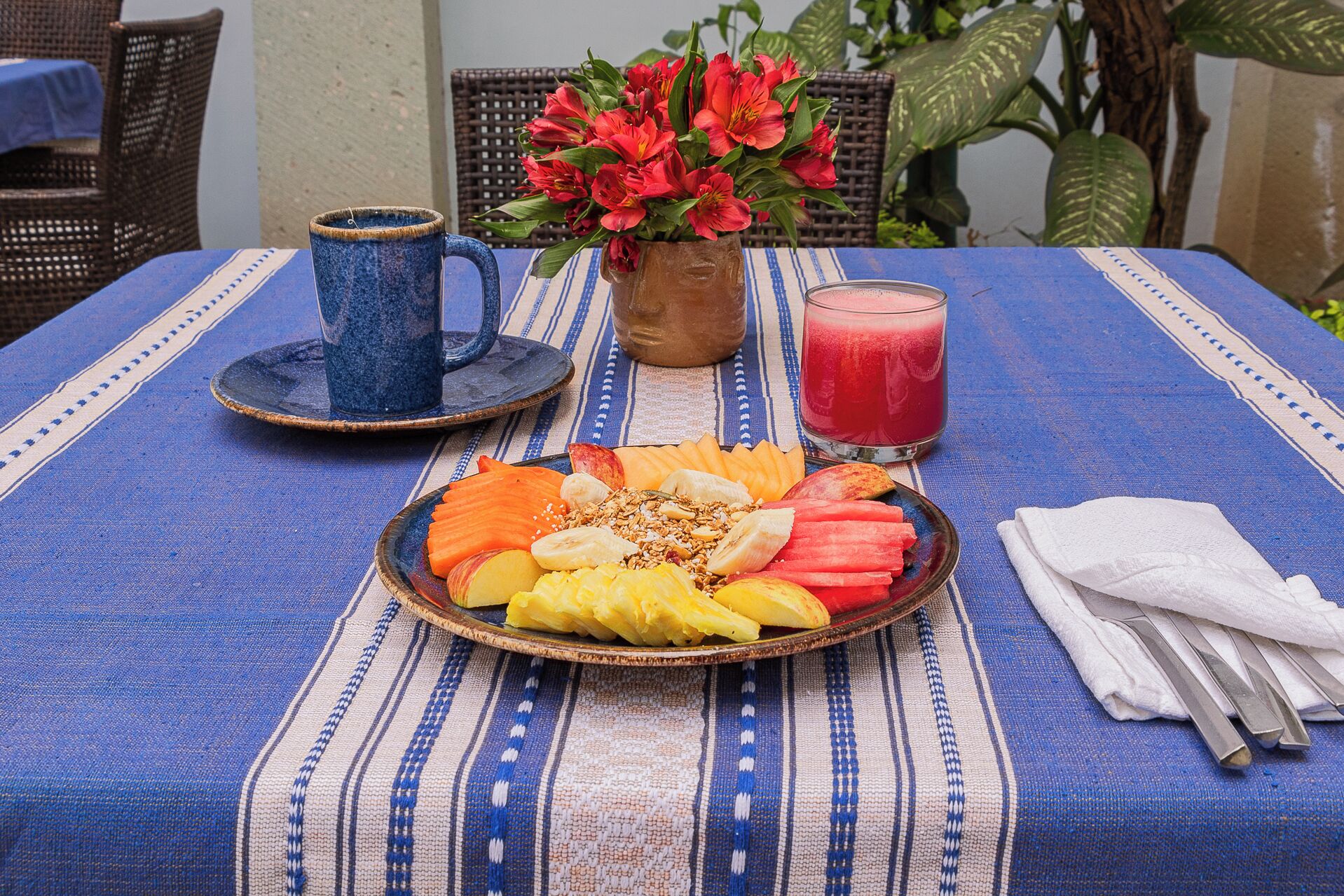 A table set with a fruit platter, a mug, a glass of red juice, and a vase of flowers.