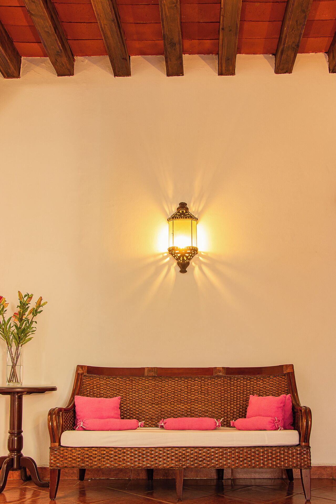 A wicker sofa with pink cushions, a small table with a vase of flowers, and a wall-mounted light fixture.