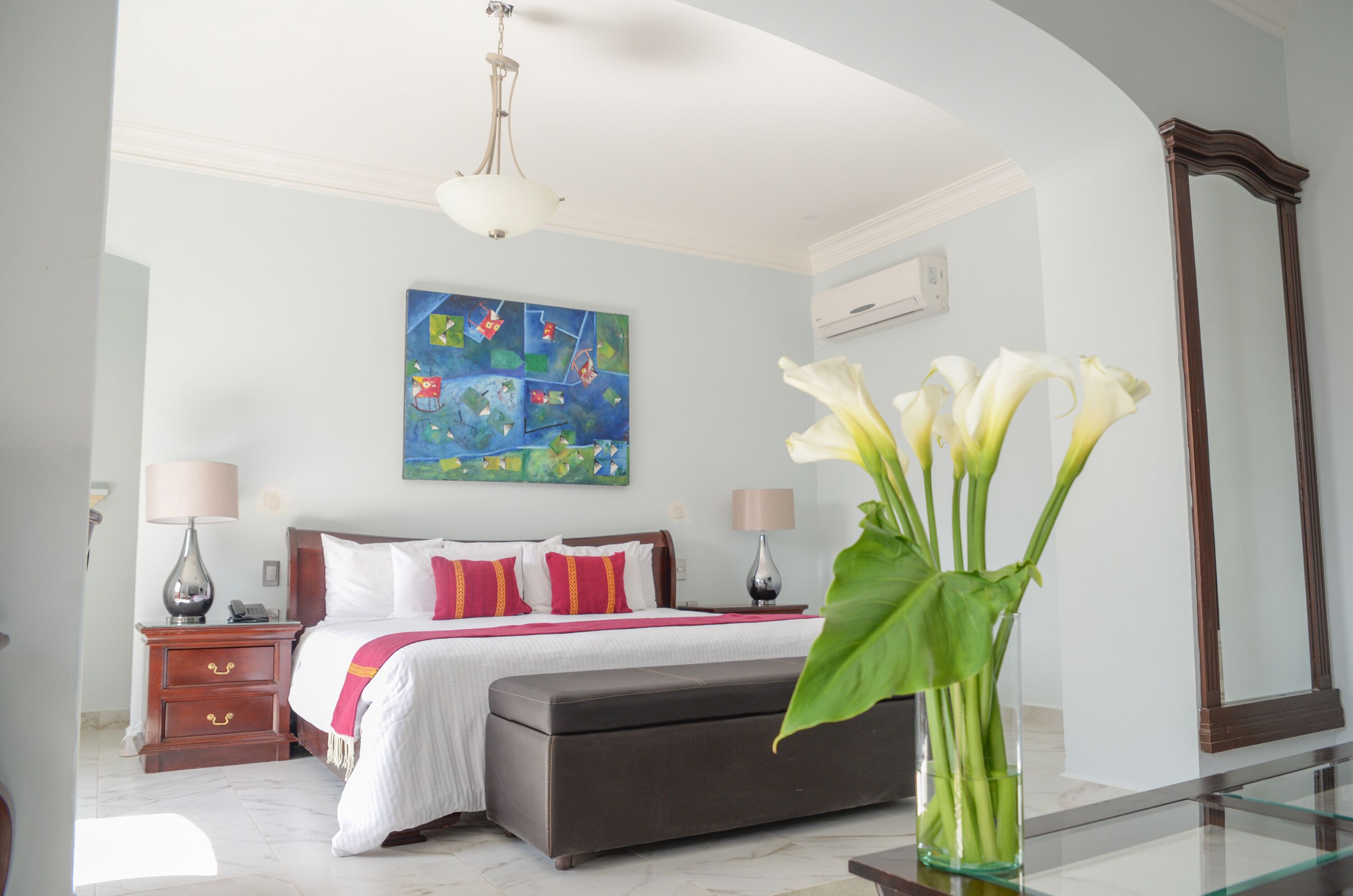 A hotel room with a bed, bedside tables, a nightstand, a large mirror, and a painting on the wall.