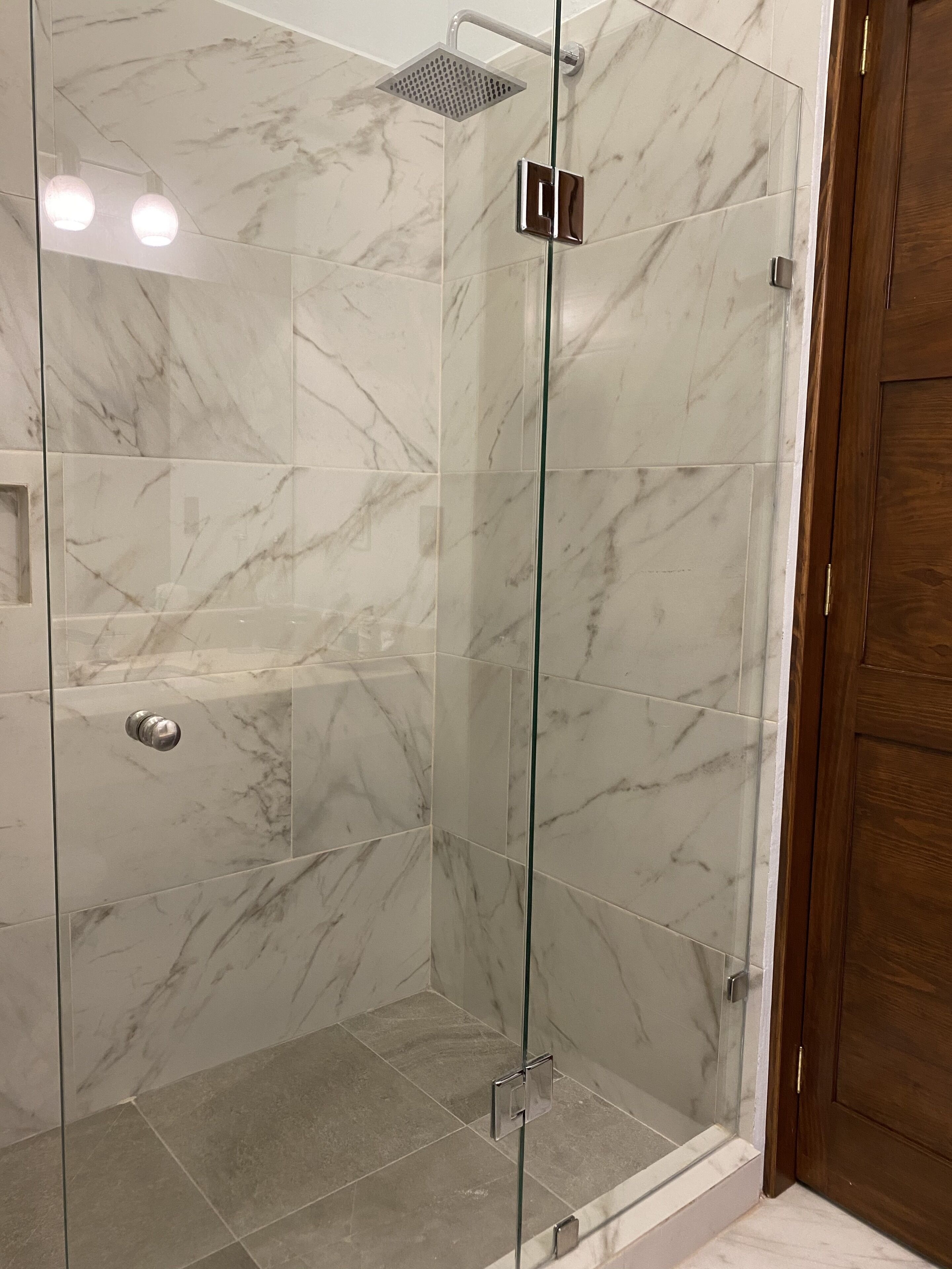 Ducha con paredes de vidrio, cabezal de ducha manual, azulejos de mármol y puerta de madera.