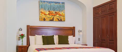 A bedroom with a wooden headboard, a large bed with white bedding, a colorful throw blanket, a nightstand with a vase of flowers, and a large painting above the bed.