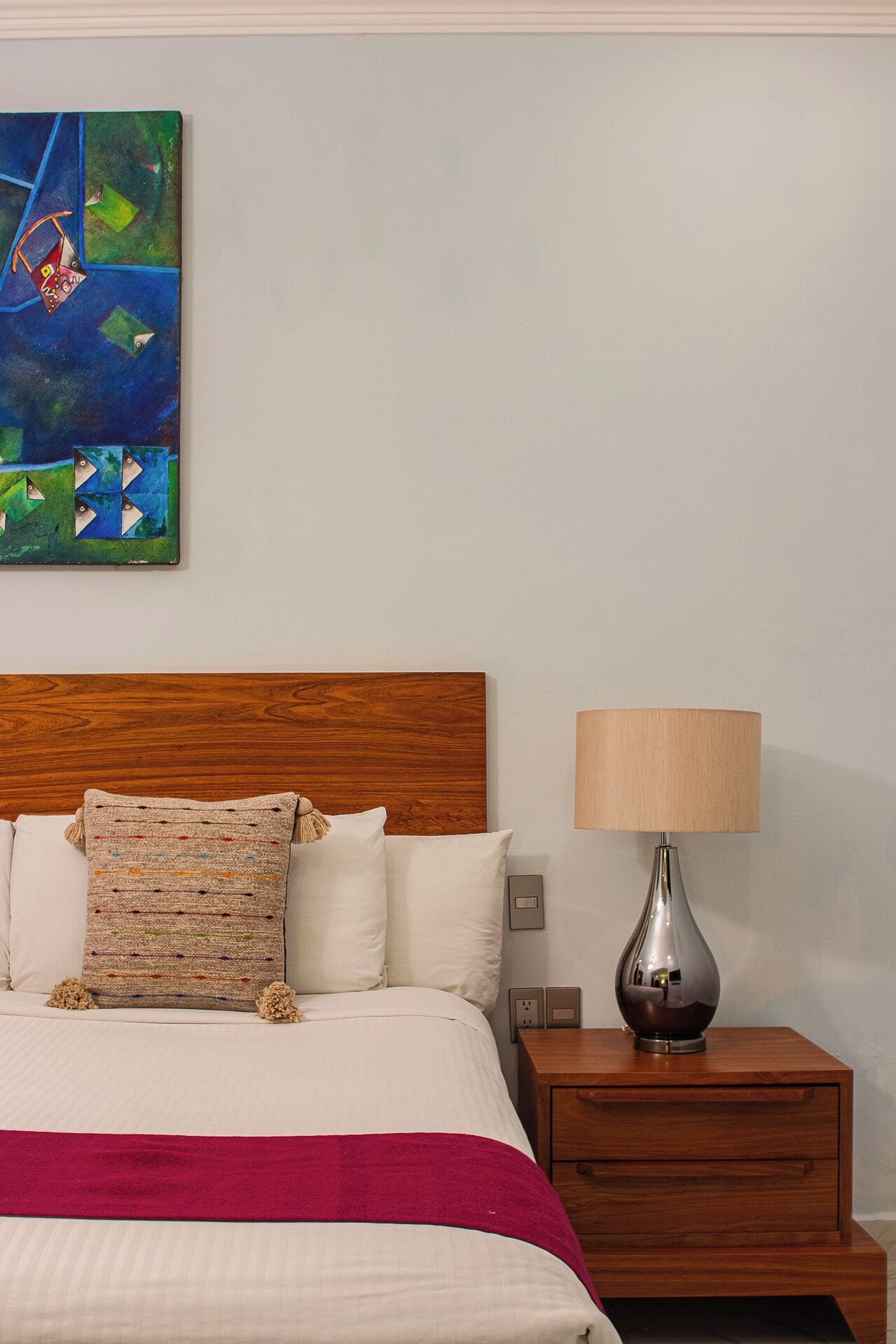 A neatly made bed with a wooden headboard, a colorful throw pillow, and a red blanket. A lamp and a small wooden nightstand are beside the bed.