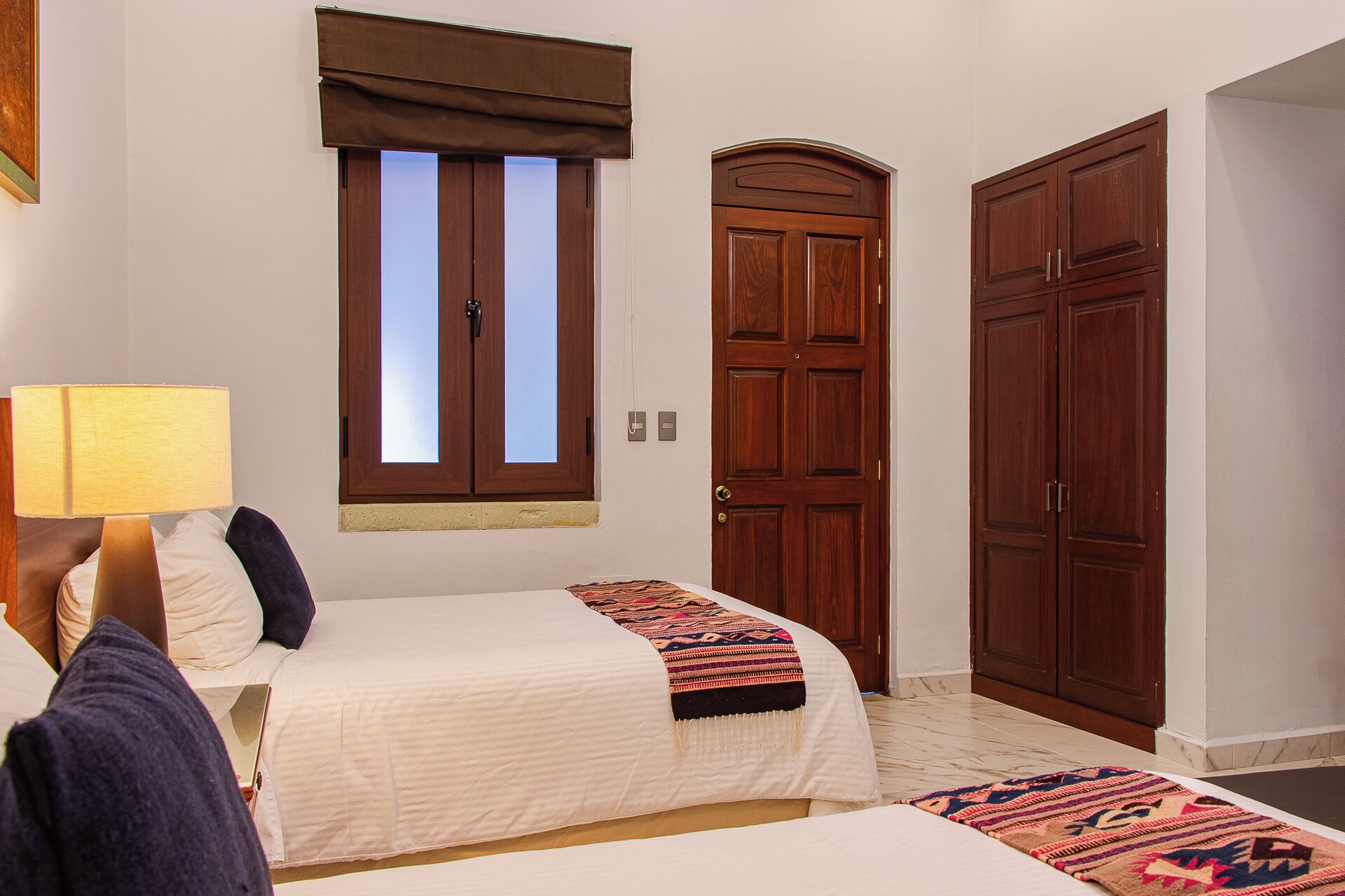 A bedroom with two beds, a window with blinds, a door, and a lamp.