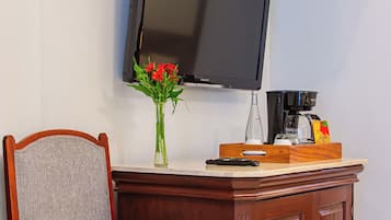 Una sedia in legno con seduta e schienale imbottiti, una TV a schermo piatto montata a parete, un piccolo tavolino con un vaso di fiori, una macchina per il caffè e un telecomando.