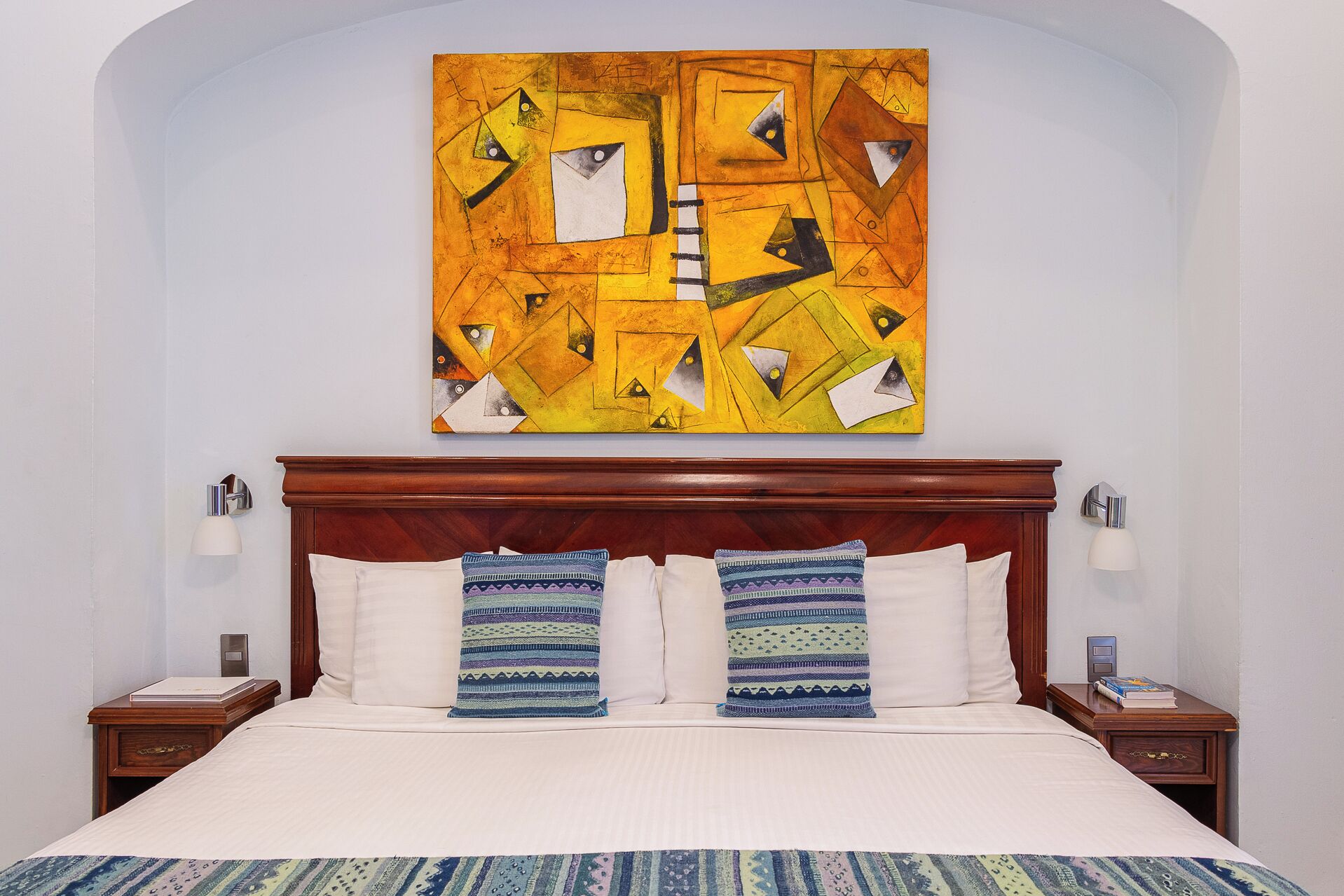 A hotel room with a wooden headboard, a bed with white linens, two patterned pillows, and a colorful abstract painting above the bed.