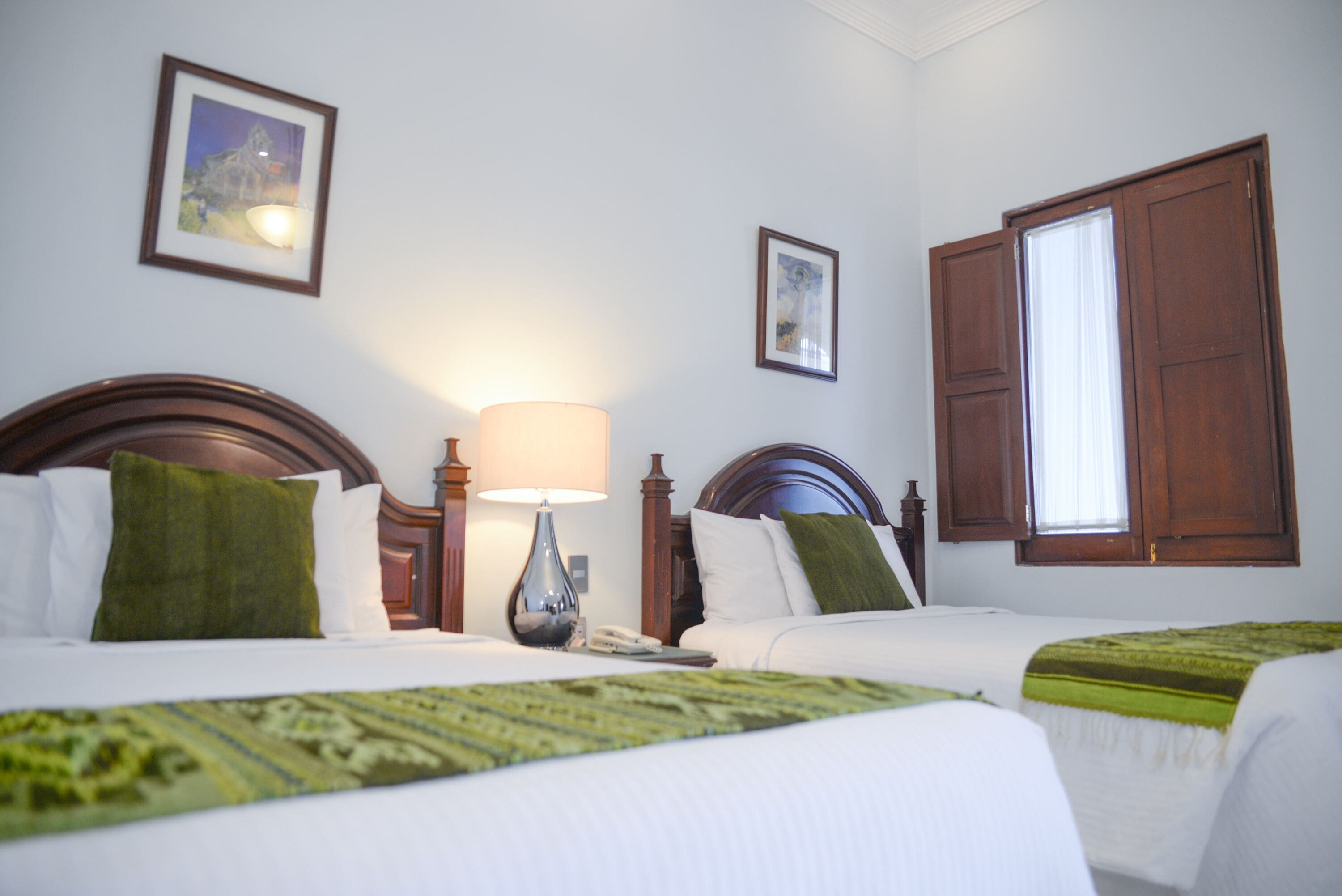 A hotel room with two beds, a nightstand with a lamp, and a window with wooden shutters.