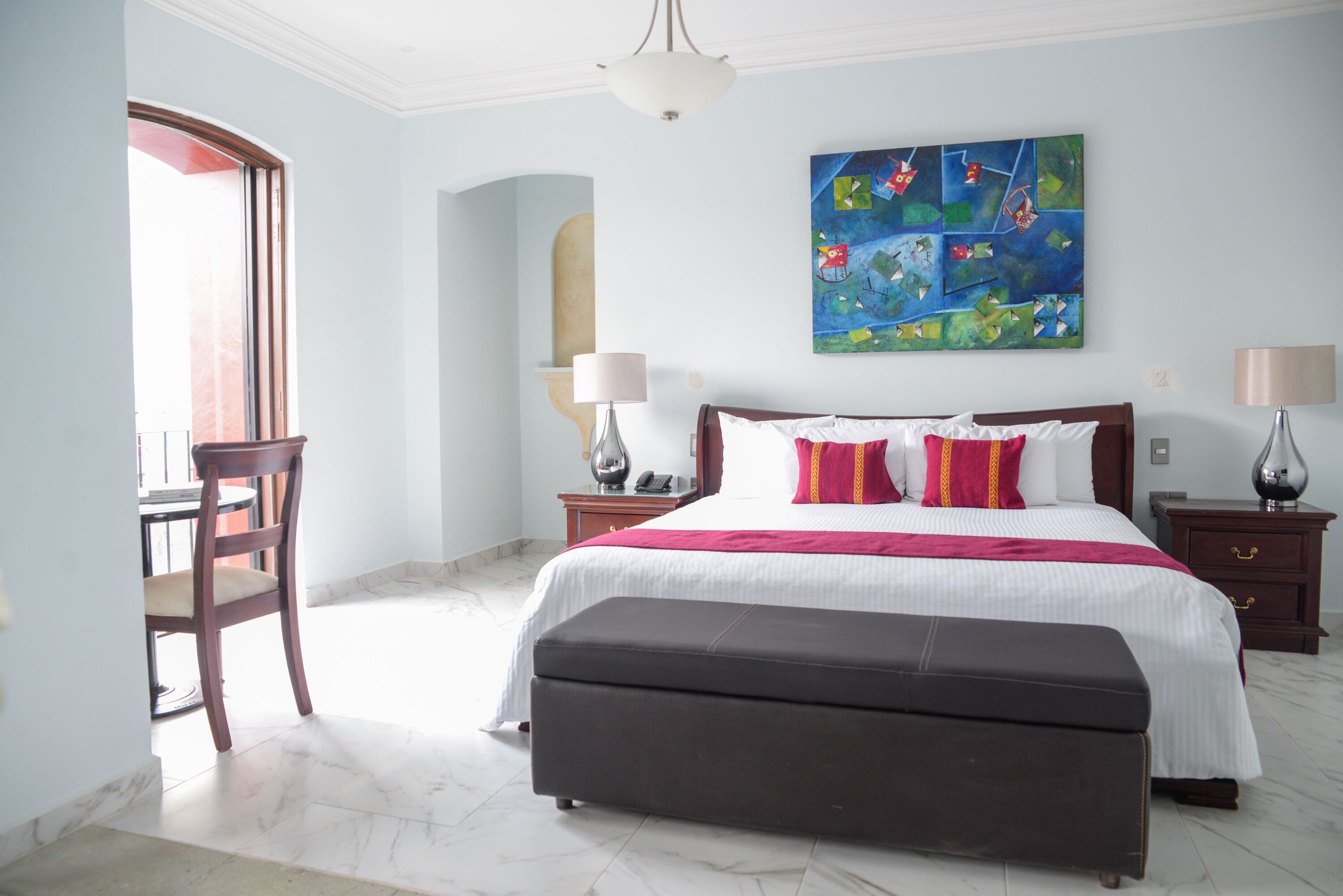 A bedroom with a large bed, a desk, and a chair. There is a painting on the wall and a window with a view.