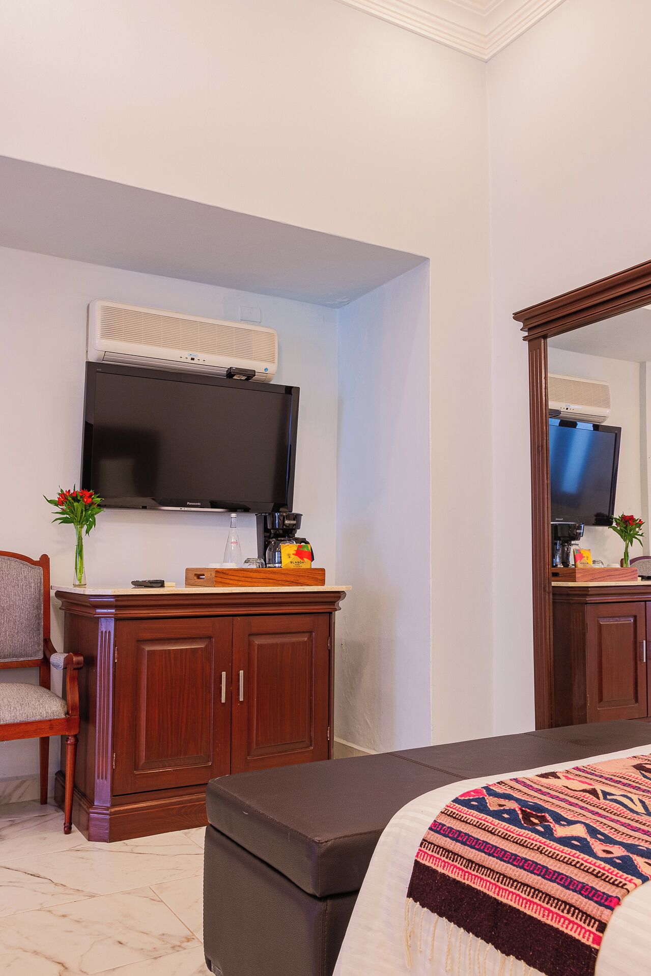 A hotel room with a bed, a TV mounted on the wall, a wooden dresser, a coffee maker, and a vase with flowers.