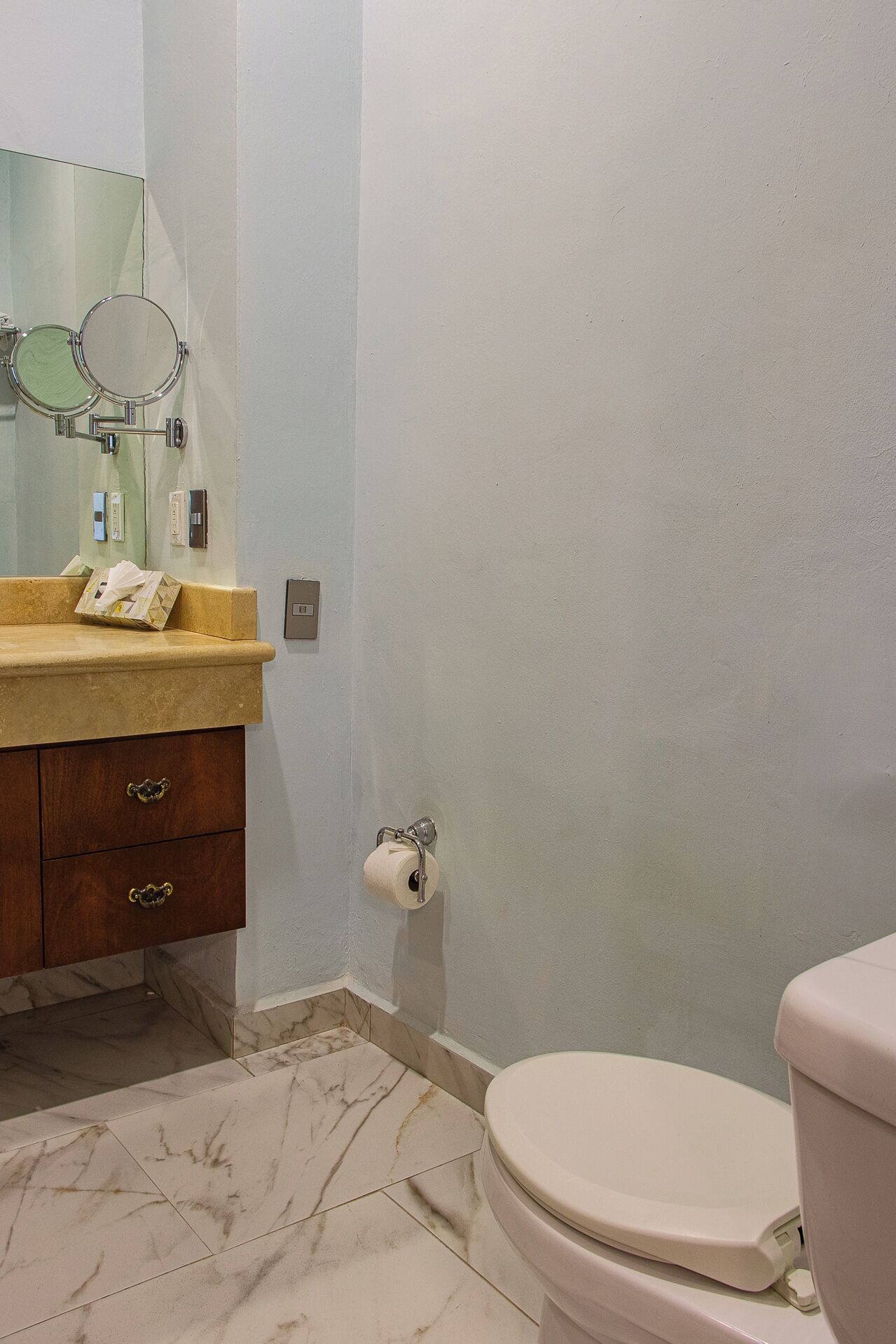 A bathroom with a marble-tiled floor, a beige toilet, a wooden vanity with a mirror, and a toilet paper holder.