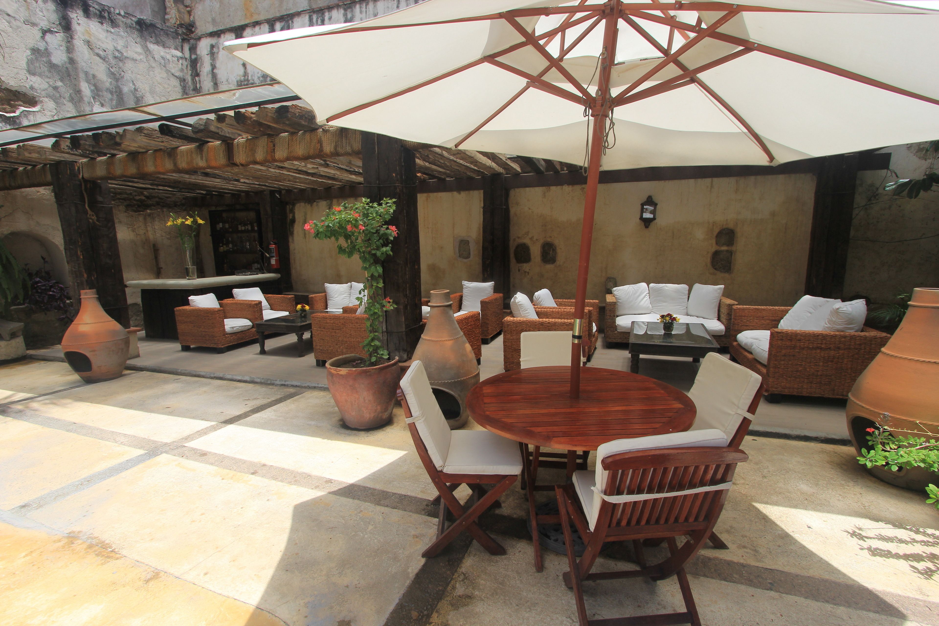 An outdoor patio area with a round wooden table, white cushioned chairs, and a large umbrella. There are wicker sofas and potted plants around.