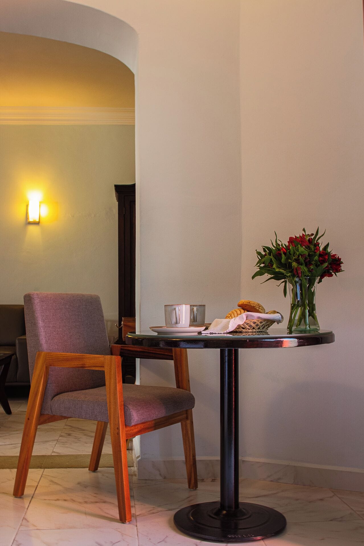 Un coin repas avec une table ronde, une chaise, un vase avec des fleurs rouges et un luminaire fixé au mur.