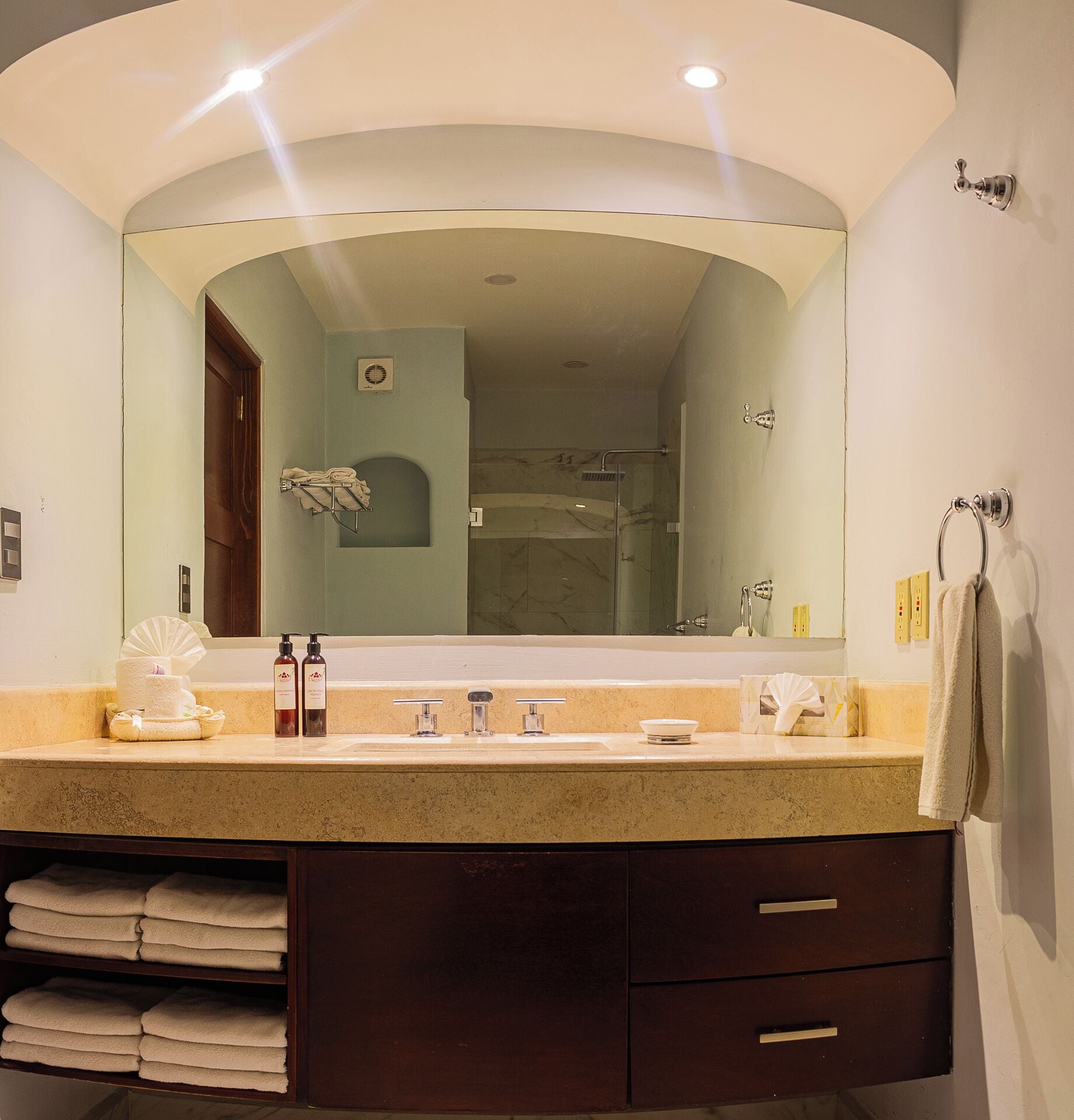 A bathroom with a large mirror, a sink, and a shower. There are towels and toiletries on the counter.