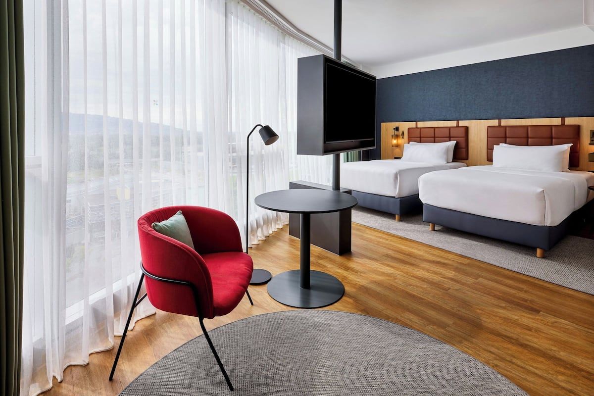 Une chambre d’hôtel avec deux lits, un téléviseur, un fauteuil rouge et une vue sur la ville à travers des rideaux transparents.